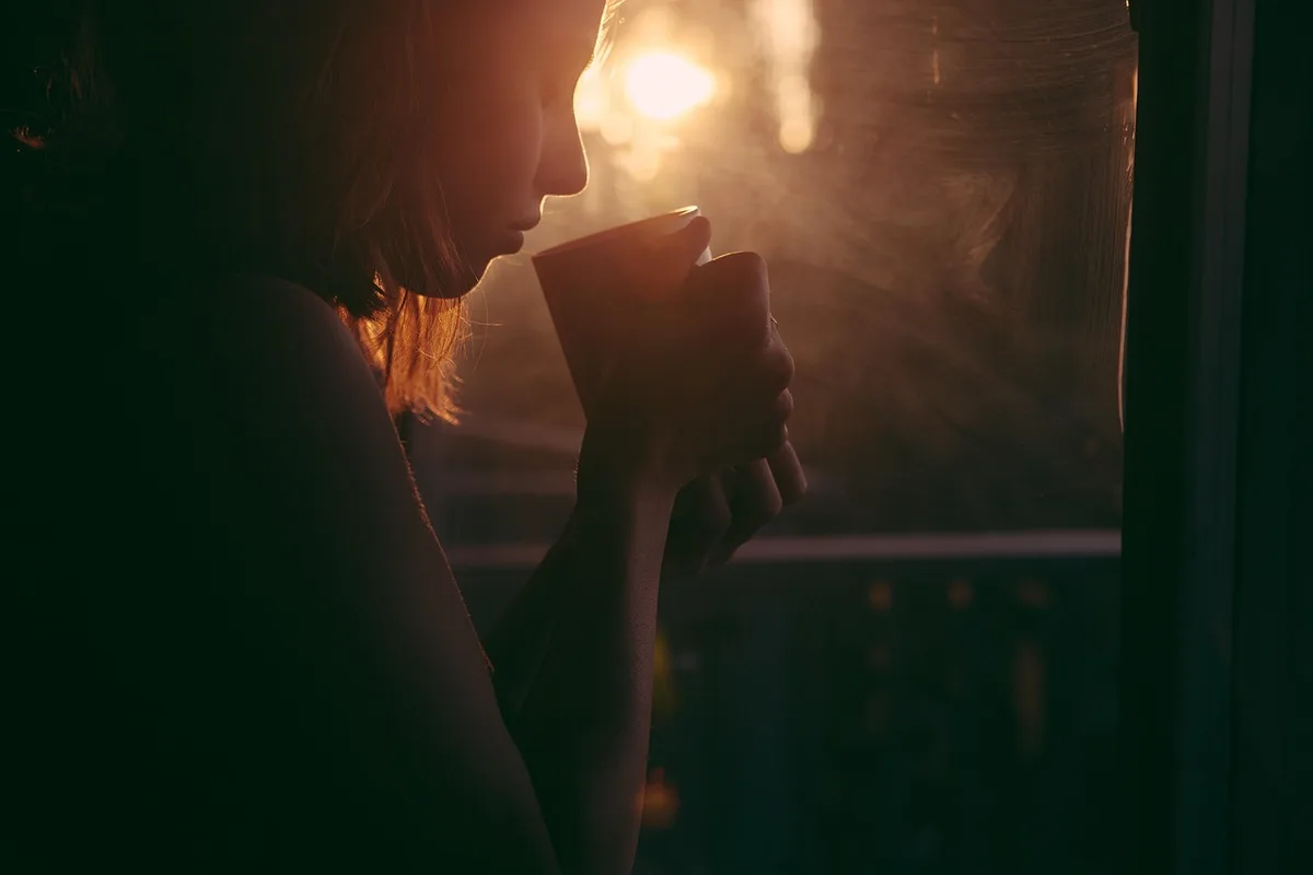 Mains anonymes tenant une tasse près d'une fenêtre, mouchoirs sur la table, lumière douce